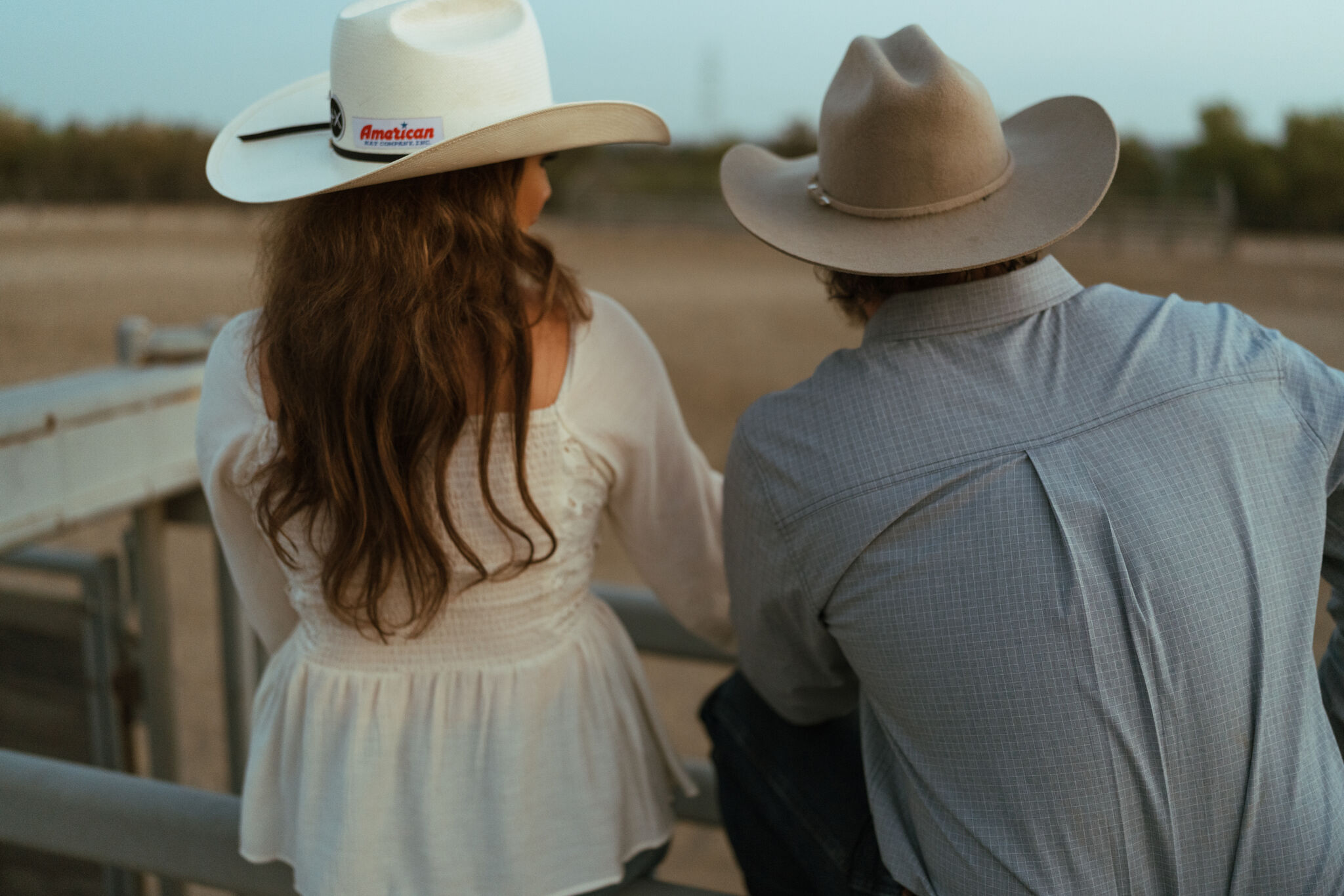 Couple in cowboy hats sitting close together on an arena fence at dusk