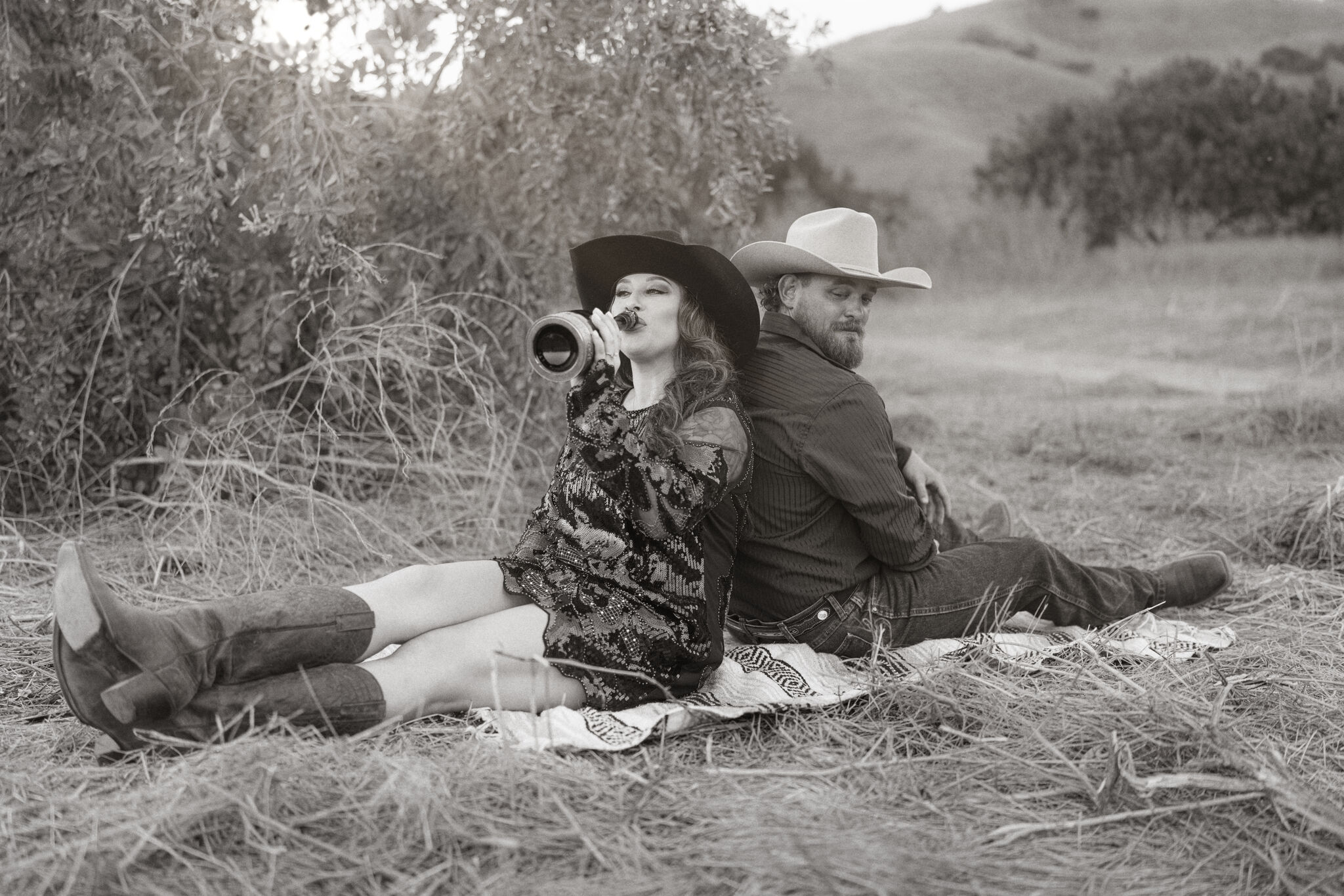 Couple in cowboy hats sitting back to back in a hay field, sharing champagne